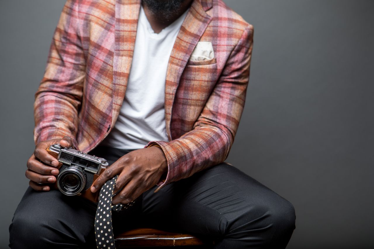 services-img A fashionable man in a plaid blazer holds a vintage camera, seated indoors.