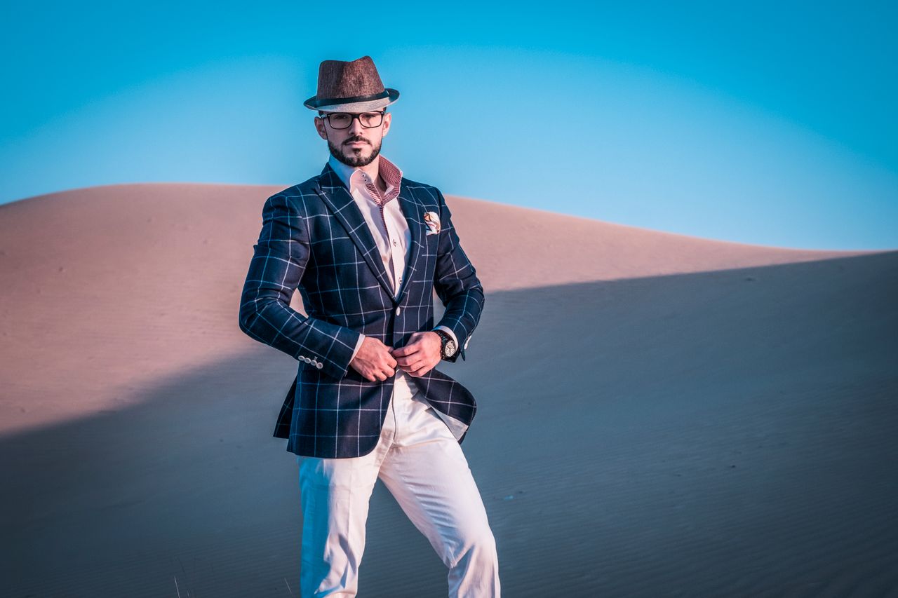 services-02 Fashionable man posing in desert in checkered blazer and fedora under clear sky.